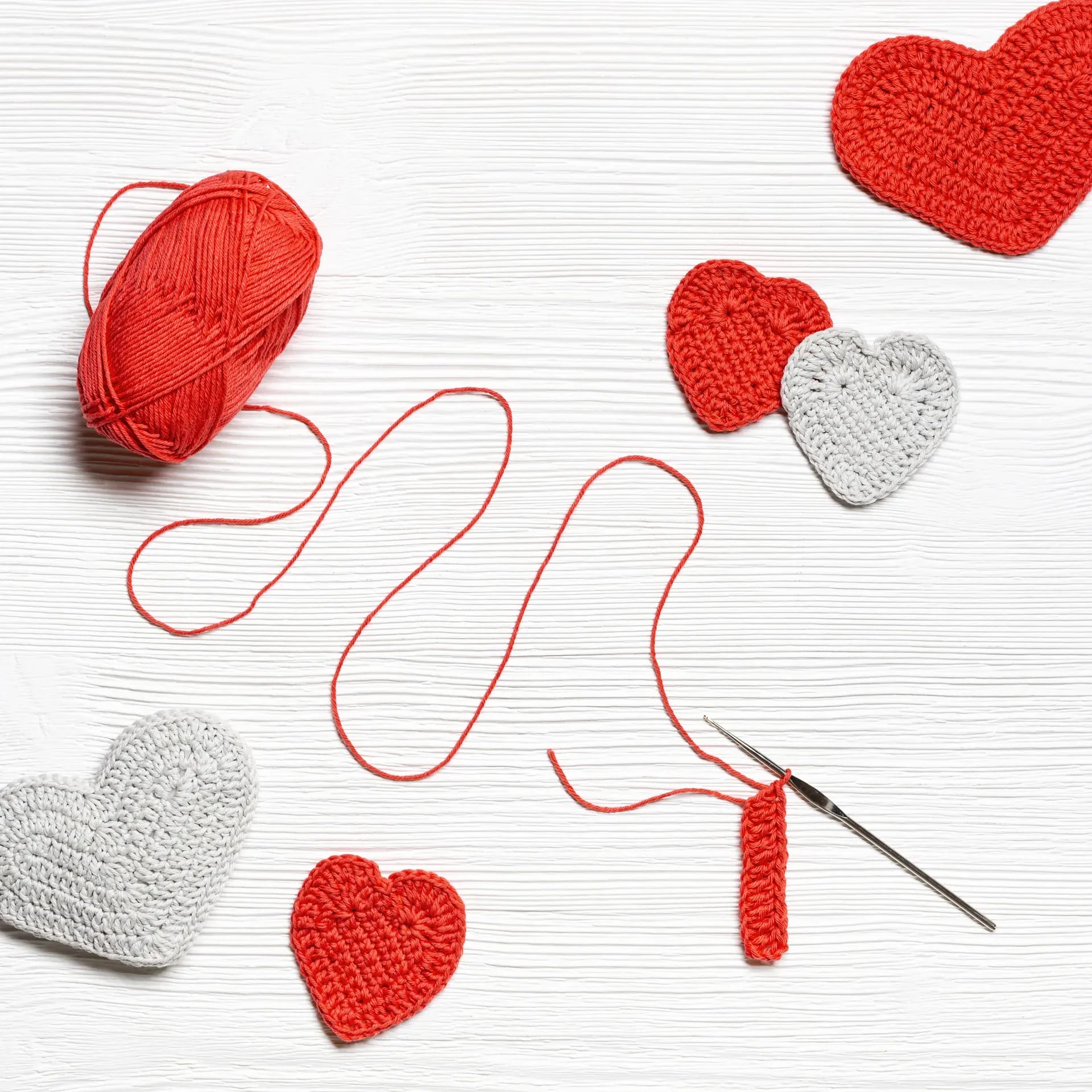 Cœurs au crochet rouges et gris, une pelote rouge et un crochet sur une surface en bois blanc